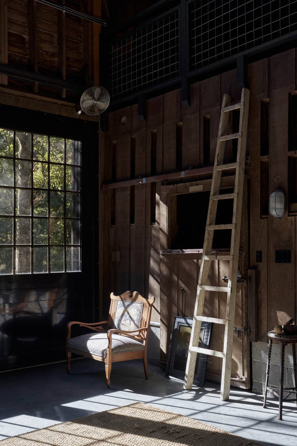 Sunlit barn interior corner with antique armchair, leaning wooden ladder, and original horse stall doors
