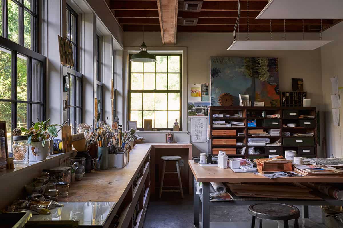 Artist studio with wall of steel-framed windows, paint-covered workbench, brushes, and large canvas in progress