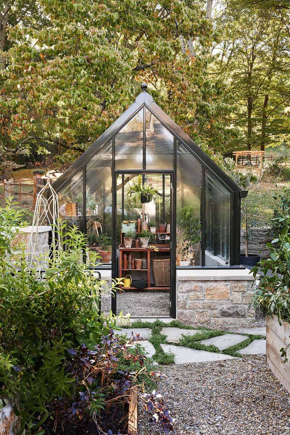 Small black steel-framed greenhouse with gabled roof on stone base, filled with potted plants and garden tools