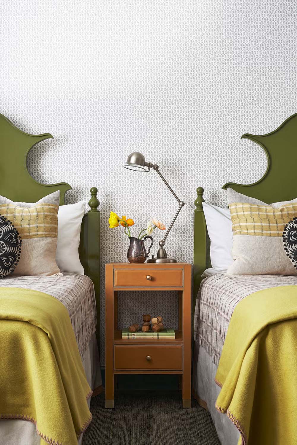 Twin bedroom with olive green carved headboards, yellow blankets, orange nightstand, and small-pattern wallpaper