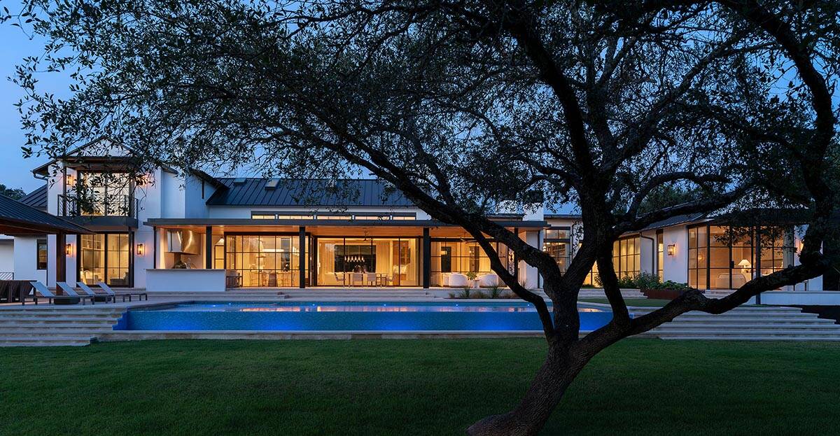 Modern farmhouse rear exterior at dusk with illuminated interiors, pool, and live oak tree