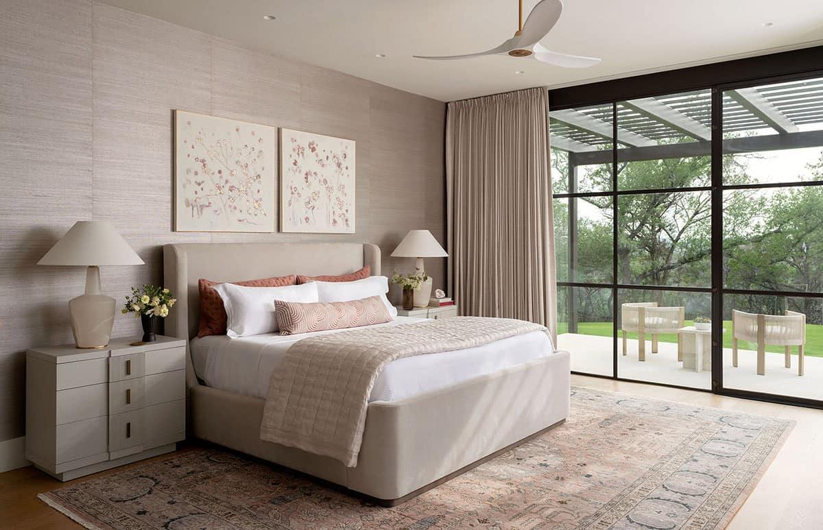 Primary bedroom with upholstered linen bed, grasscloth walls, floral artwork, and glass doors to patio