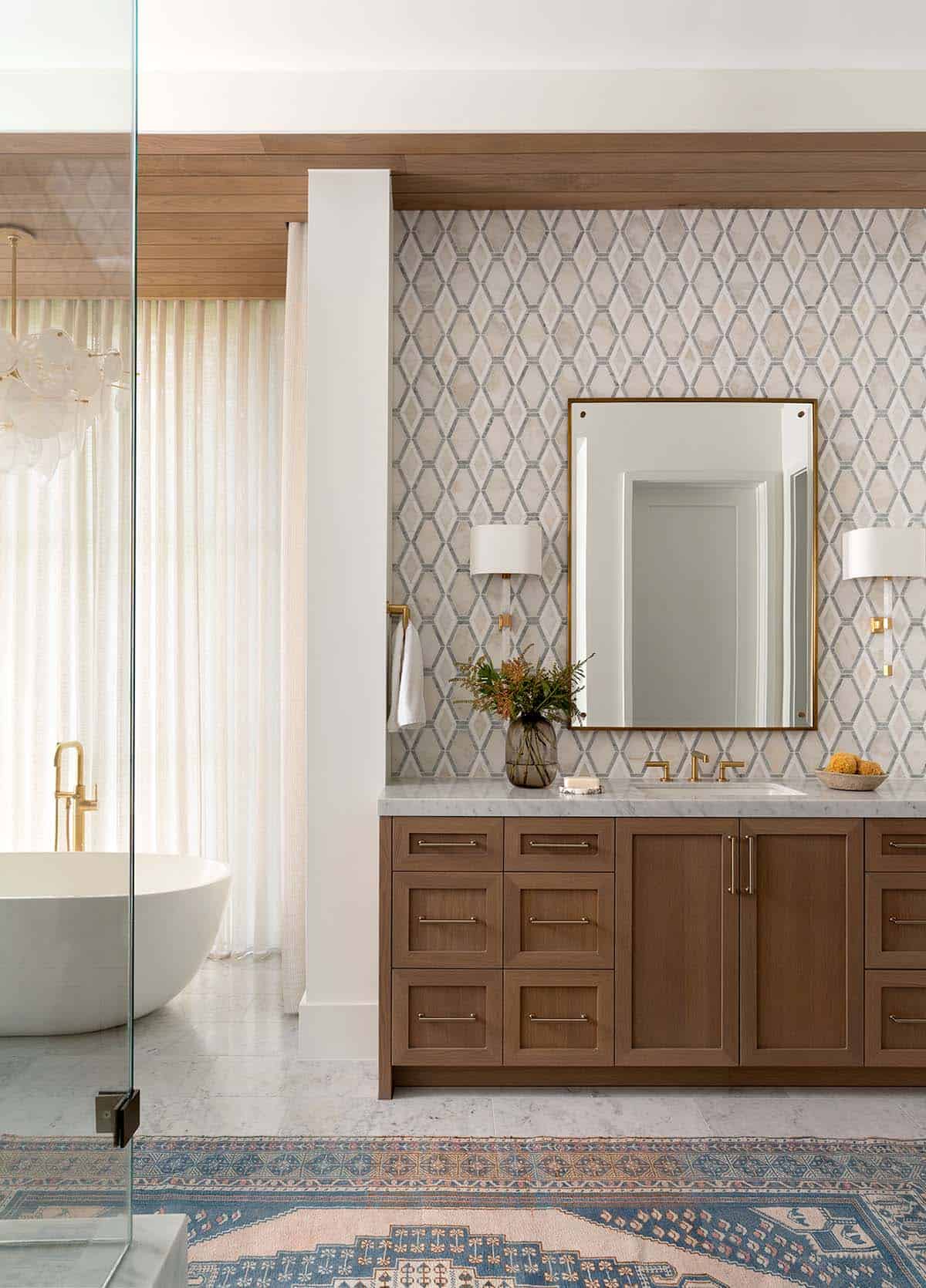 Primary bathroom vanity with walnut cabinets, diamond mosaic tile wall, brass fixtures, and freestanding soaking tub