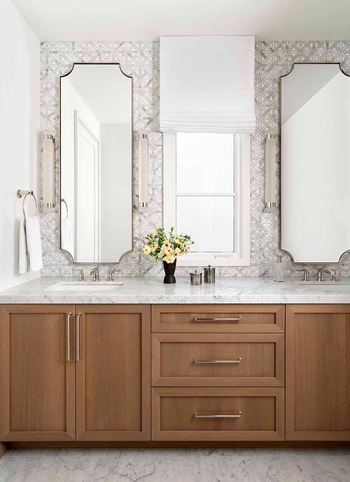Bathroom vanity with warm oak cabinets, Carrara marble countertop, geometric tile backsplash, and shaped mirrors