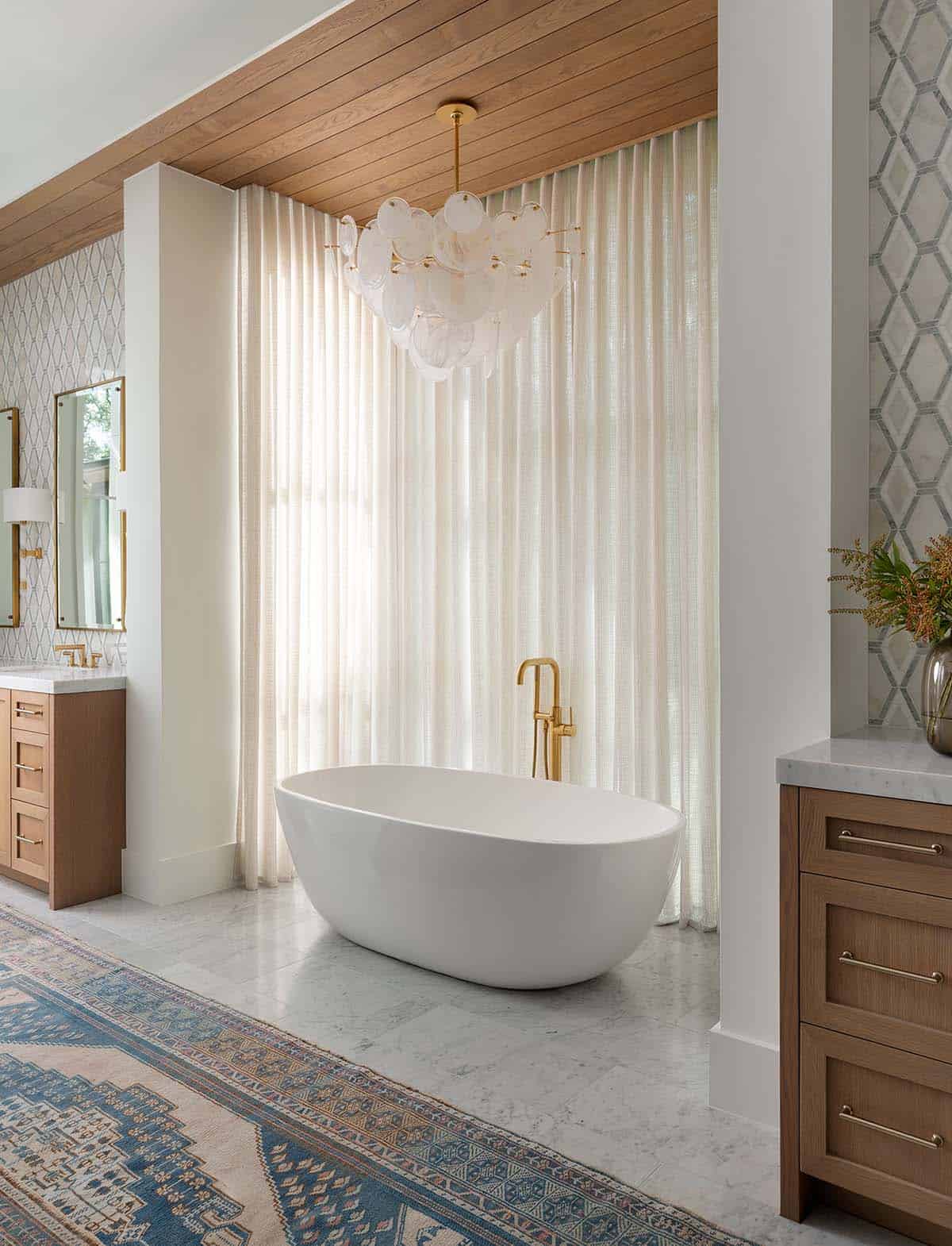 Freestanding oval soaking tub beneath sculptural glass chandelier with wood ceiling and sheer floor-length curtains