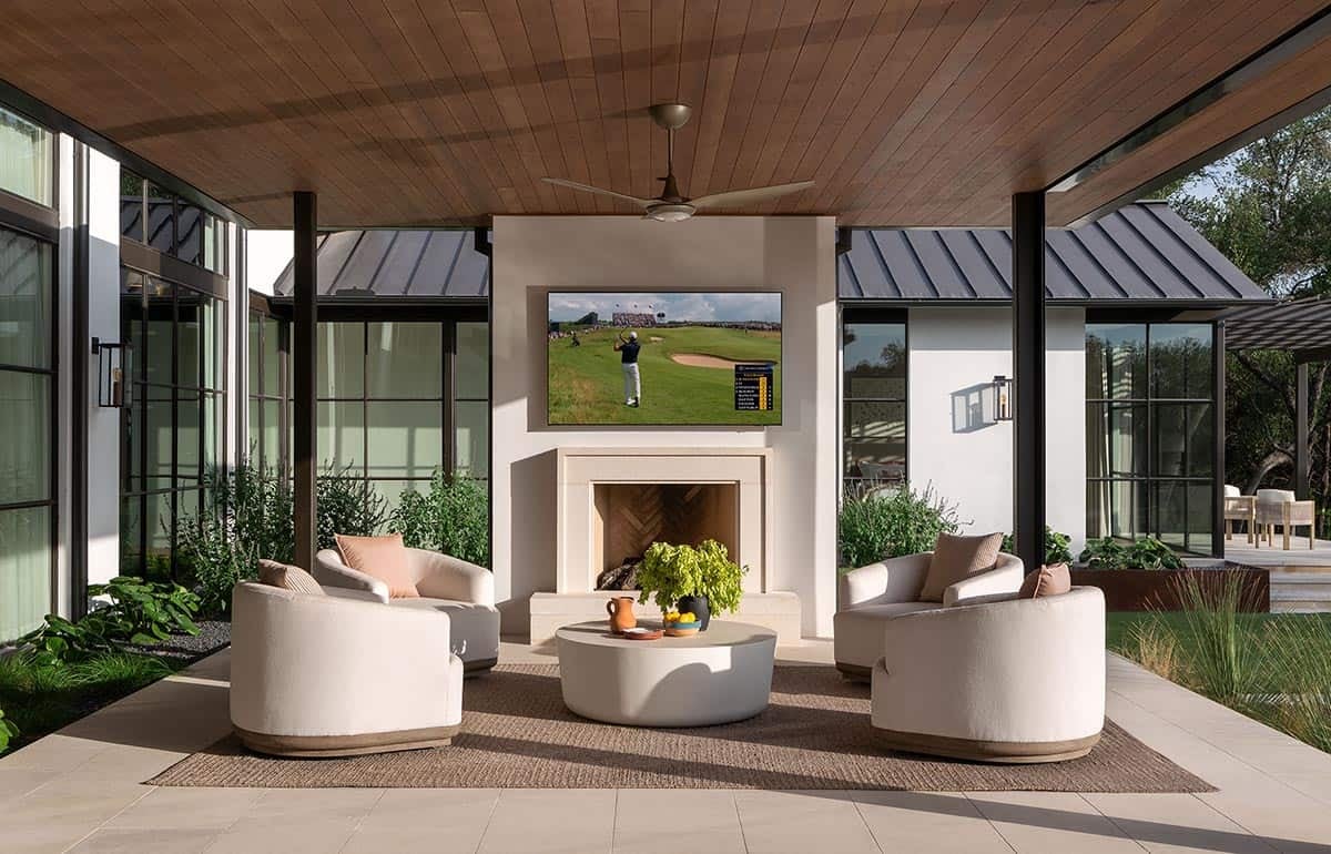 Covered outdoor living room with fireplace, TV, curved lounge chairs, wood ceiling, and steel-framed window walls