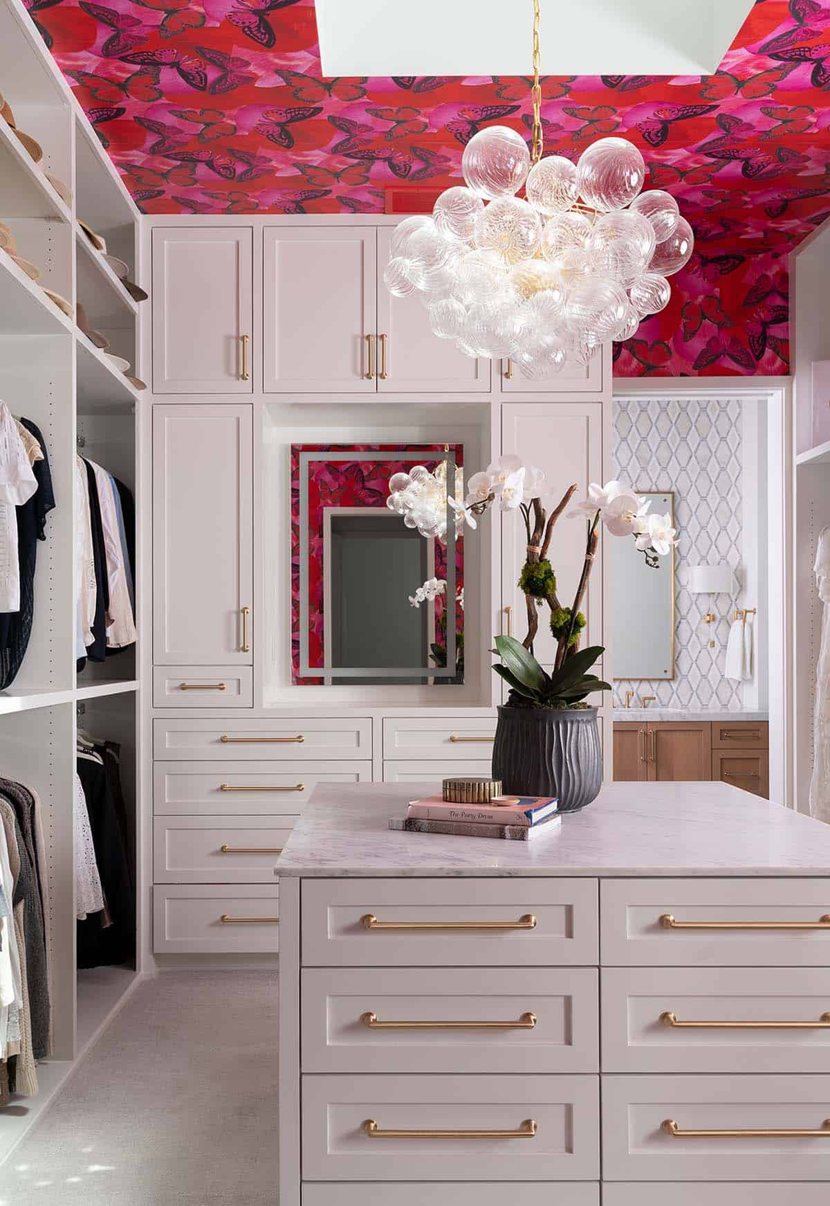 Walk-in closet with blush cabinetry, brass hardware, bubble chandelier, and bold butterfly-print ceiling wallpaper