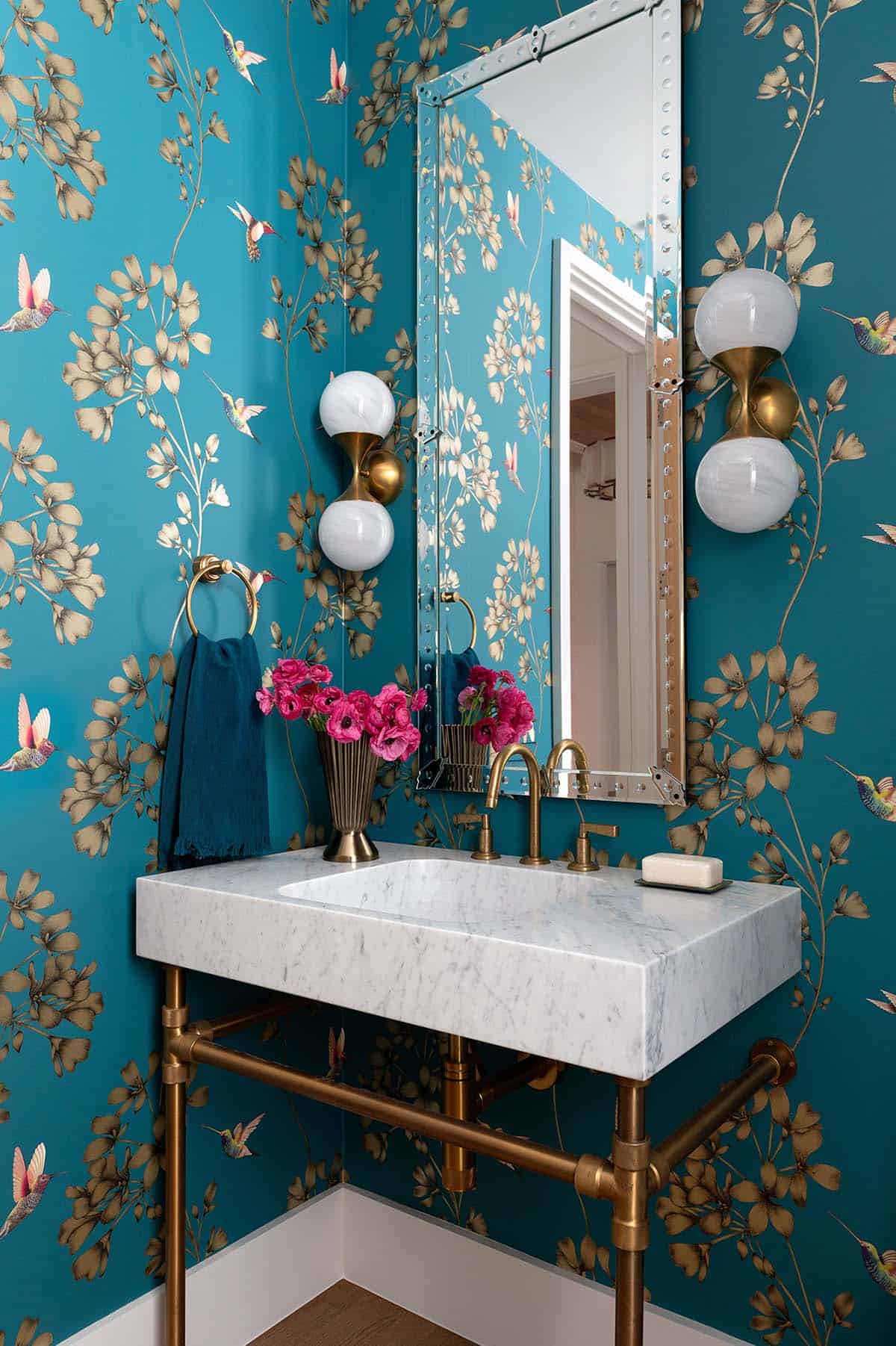 Powder room with vibrant teal hummingbird wallpaper, marble console sink on brass stand, and globe wall sconces