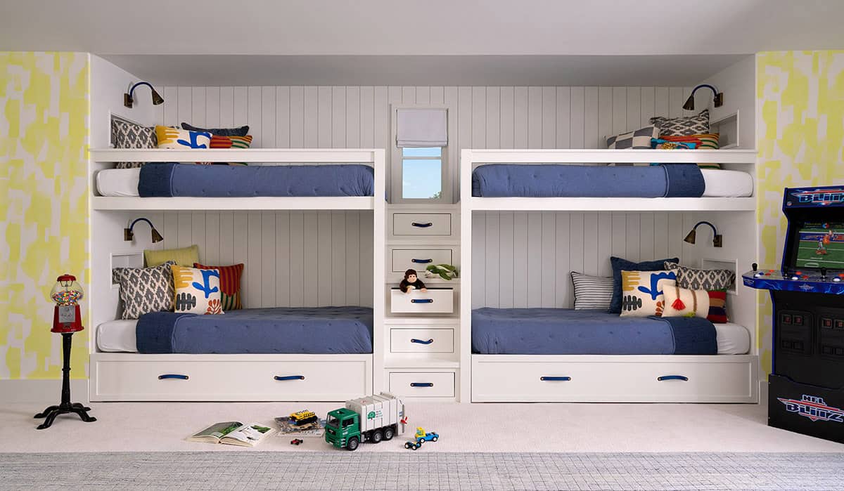Children's built-in quad bunk room with white shiplap walls, blue bedding, integrated storage drawers, and arcade game