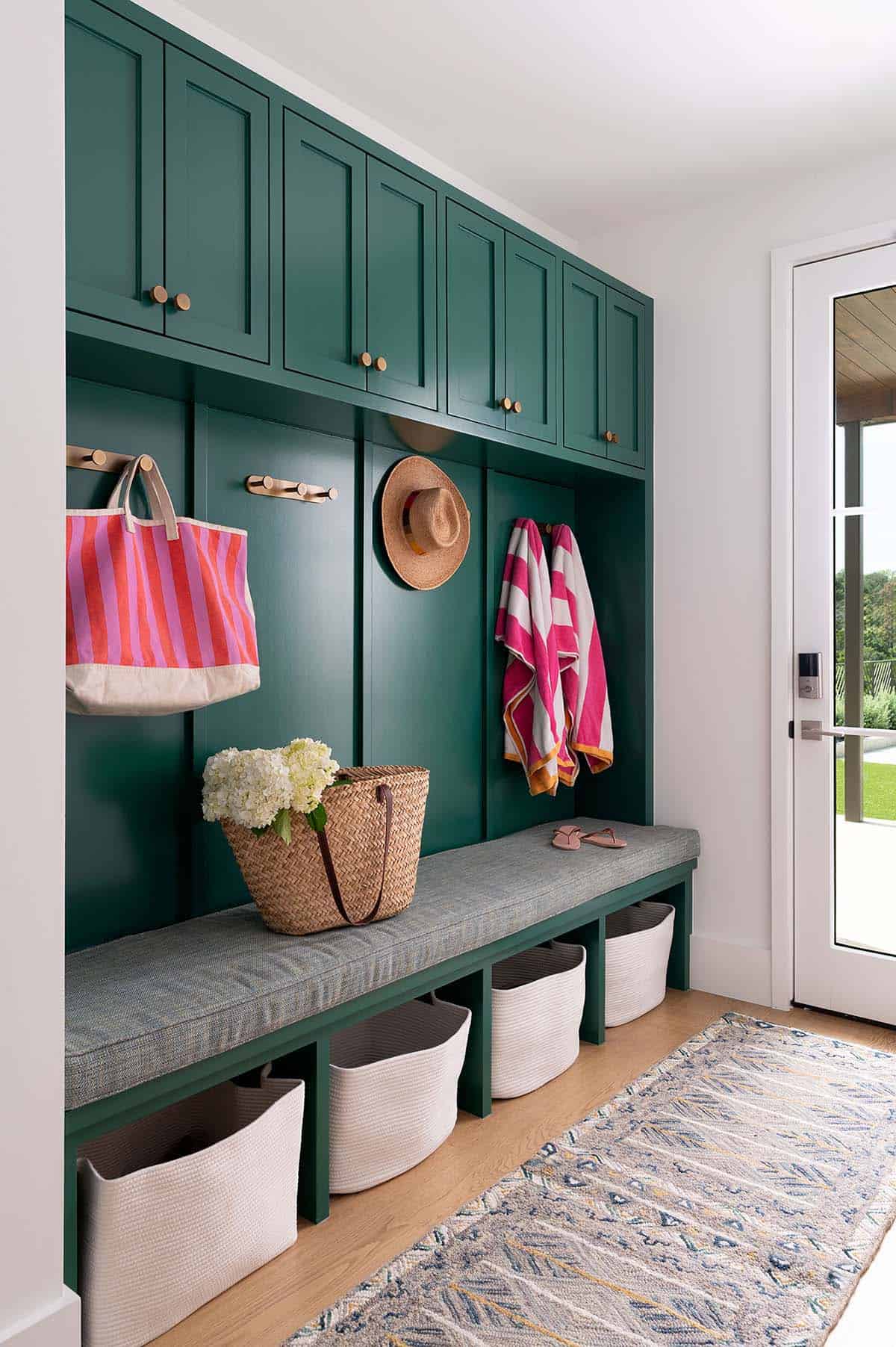 Mudroom with bold forest green built-in cabinetry, brass hooks, cushioned bench, and woven storage baskets