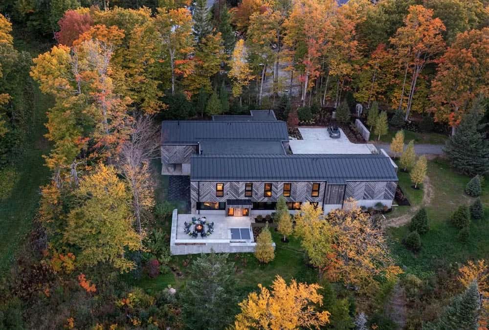 Bird's-eye drone view of ski house with standing seam metal roof and outdoor patio with fire pit amid colorful fall forest