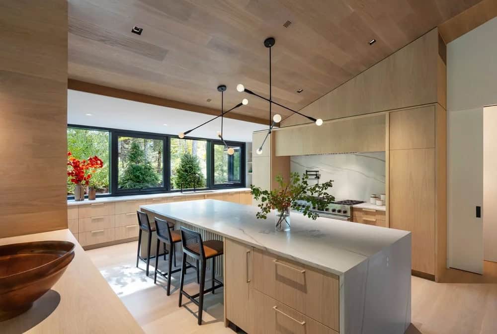 Kitchen interior with marble waterfall island, bar stools, white oak cabinets, and panoramic garden windows