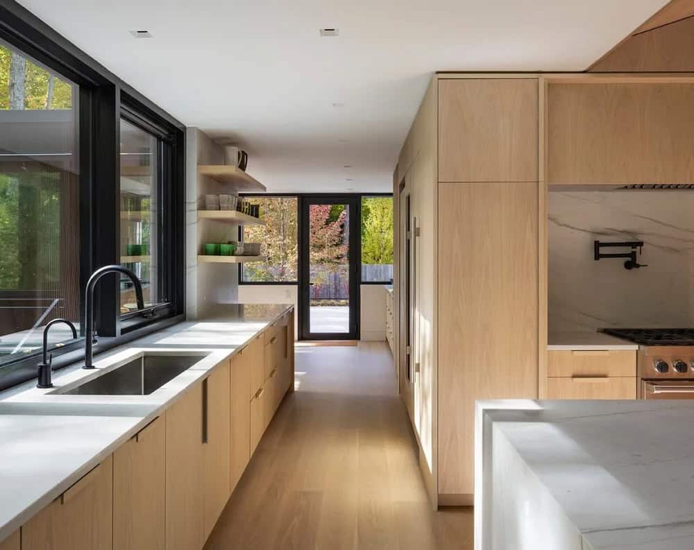 Kitchen detail showing black faucet, white quartz countertops, light wood cabinetry, and open shelving with forest views