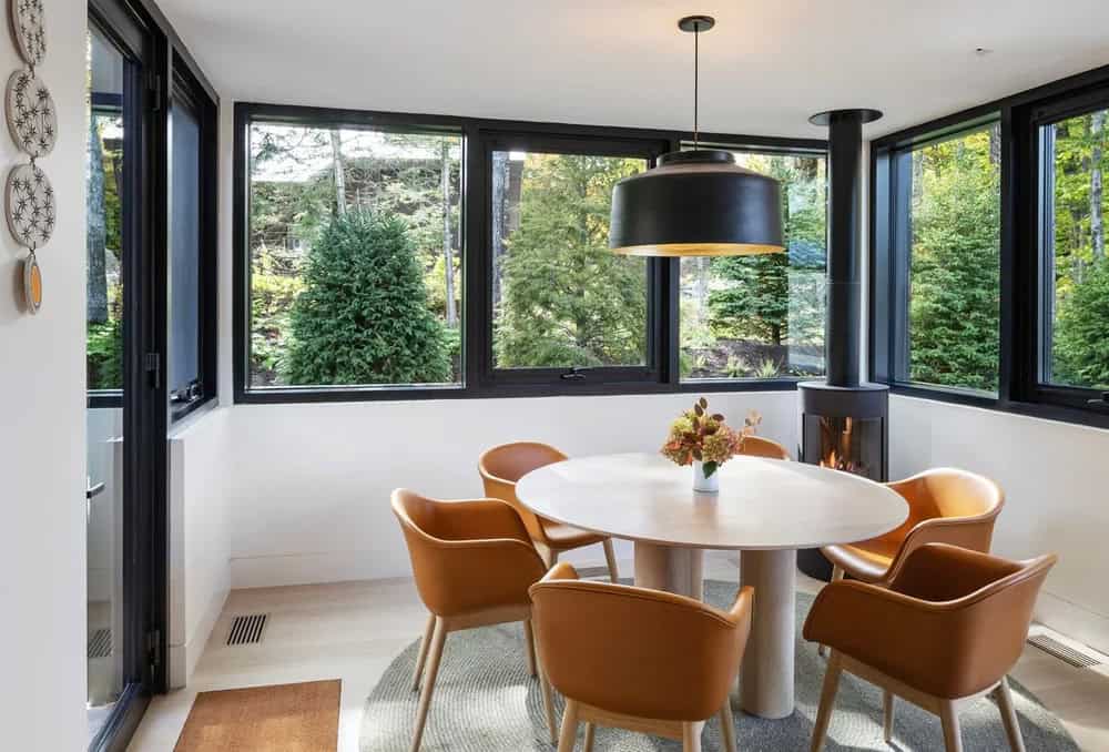 Cozy breakfast nook with round table, tan leather chairs, black wood-burning stove, and wraparound windows overlooking trees