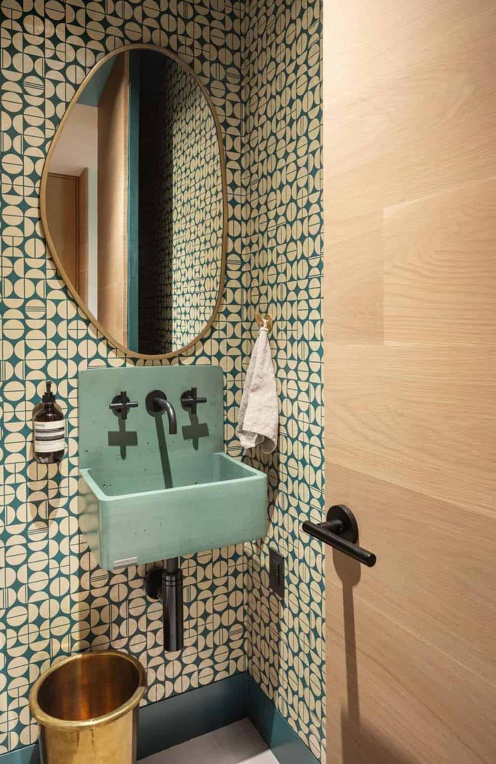 Powder room with bold teal geometric wallpaper, mint concrete wall-mounted sink, brass mirror, and matte black fixtures