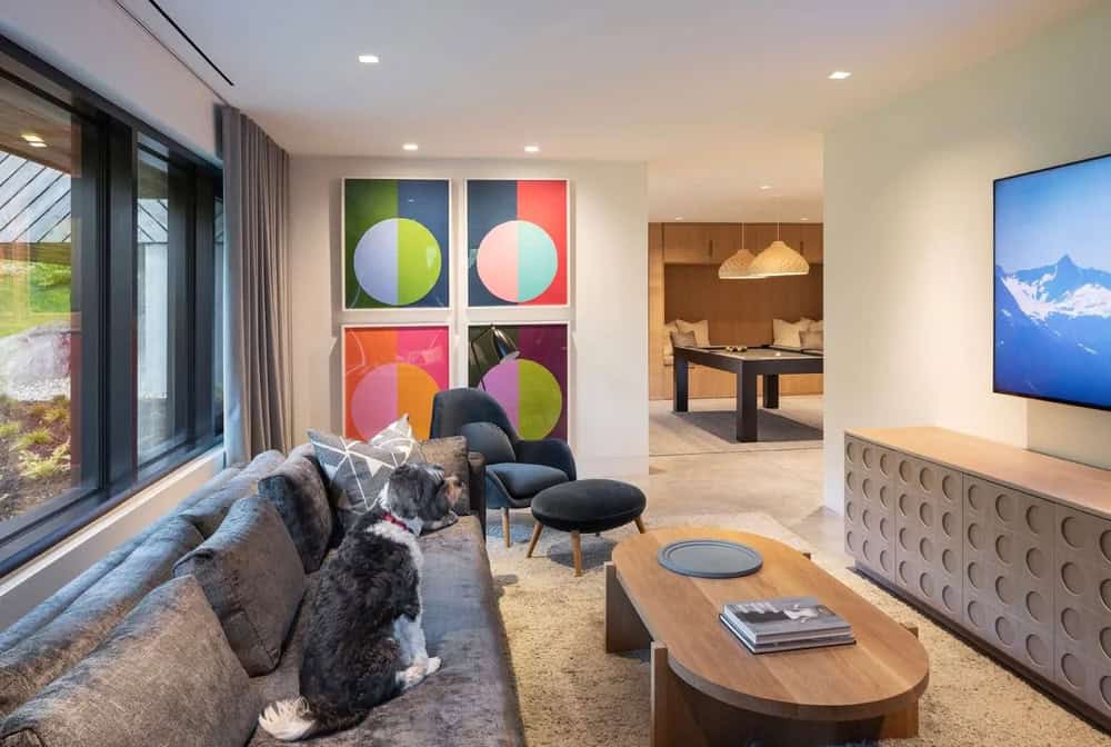 Lower level lounge with colorful circle pop art, gray sectional, dog on couch, and billiard room beyond