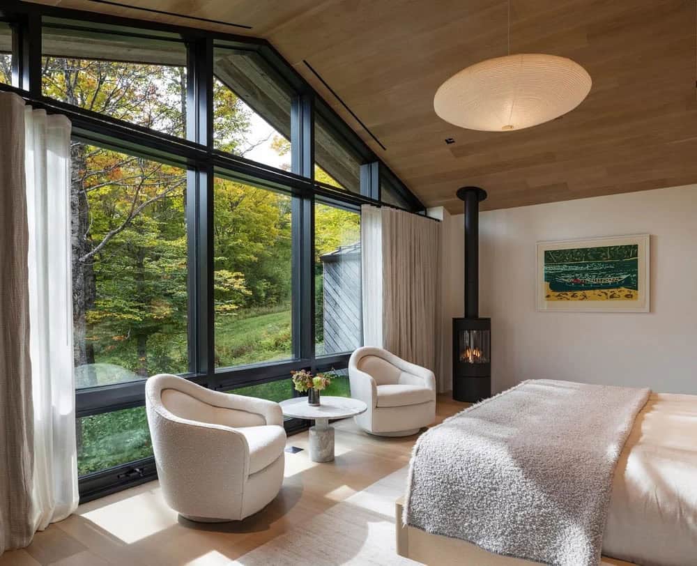 Primary bedroom sitting area with white boucl&eacute; chairs, freestanding fireplace, and gabled floor-to-ceiling forest windows
