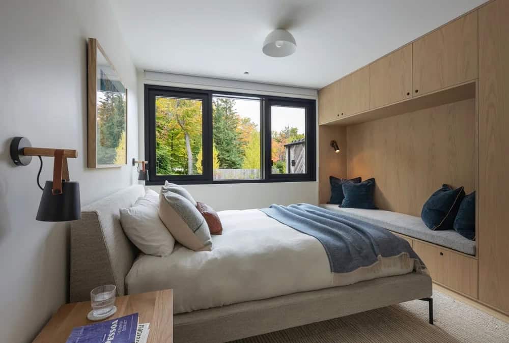 Guest bedroom with built-in oak nook, linen upholstered bed, black wall sconce, and black-framed windows overlooking autumn trees