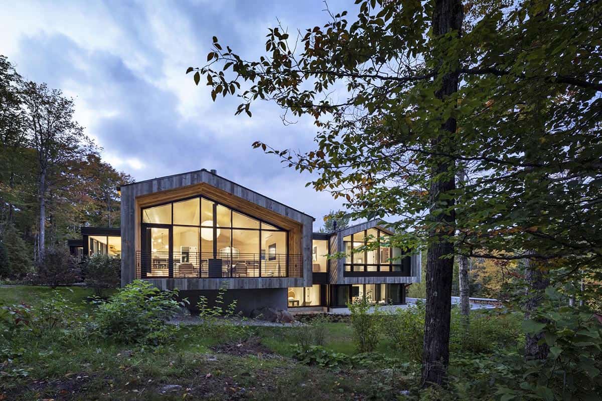 Twilight exterior of ski house with illuminated living room visible through floor-to-ceiling gabled windows