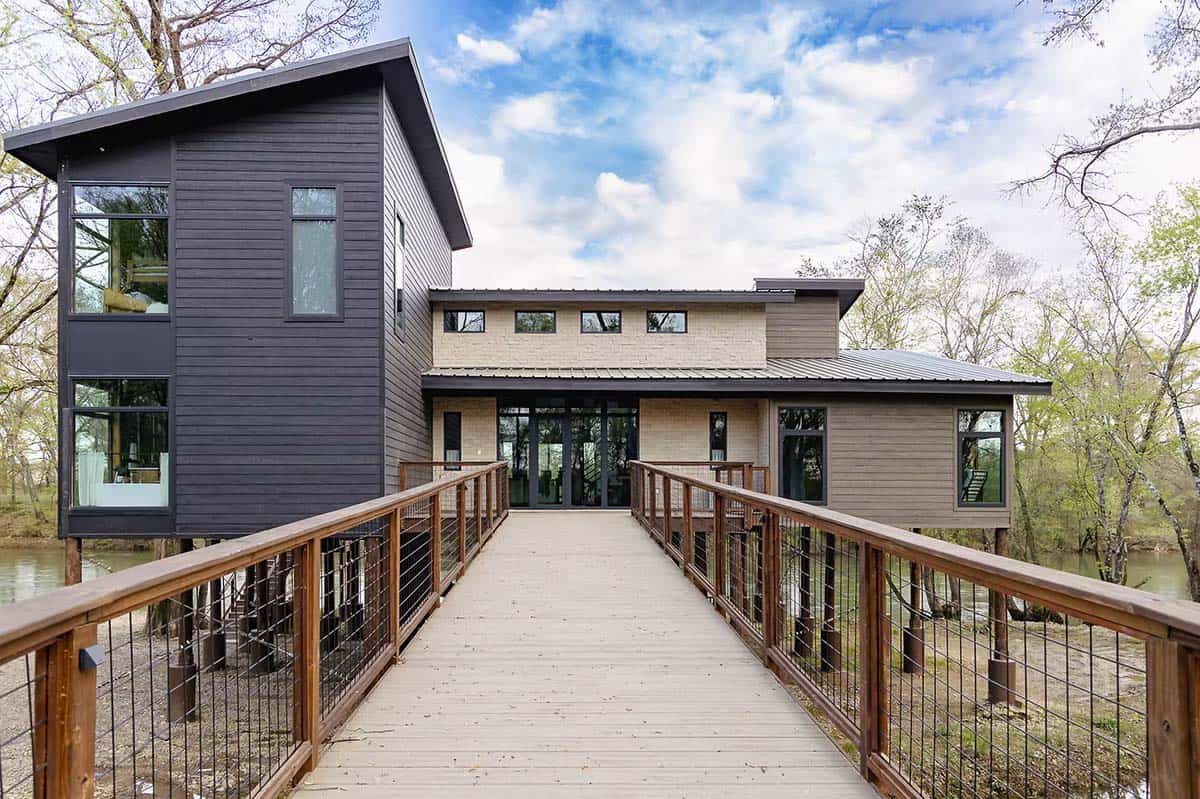 Modern rustic cabin exterior with dark and tan siding, large windows, and wooden bridge entrance
