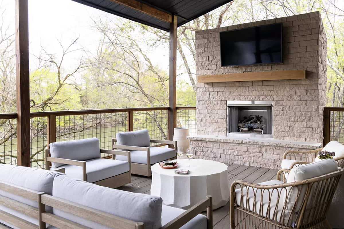 Covered outdoor porch with stone fireplace, gray lounge chairs, and wooded views