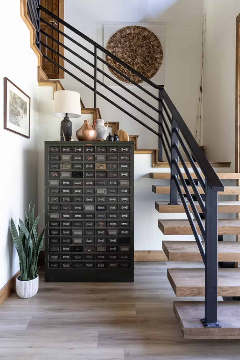 Staircase with black metal railing, vintage multi-drawer cabinet, and woven wall art