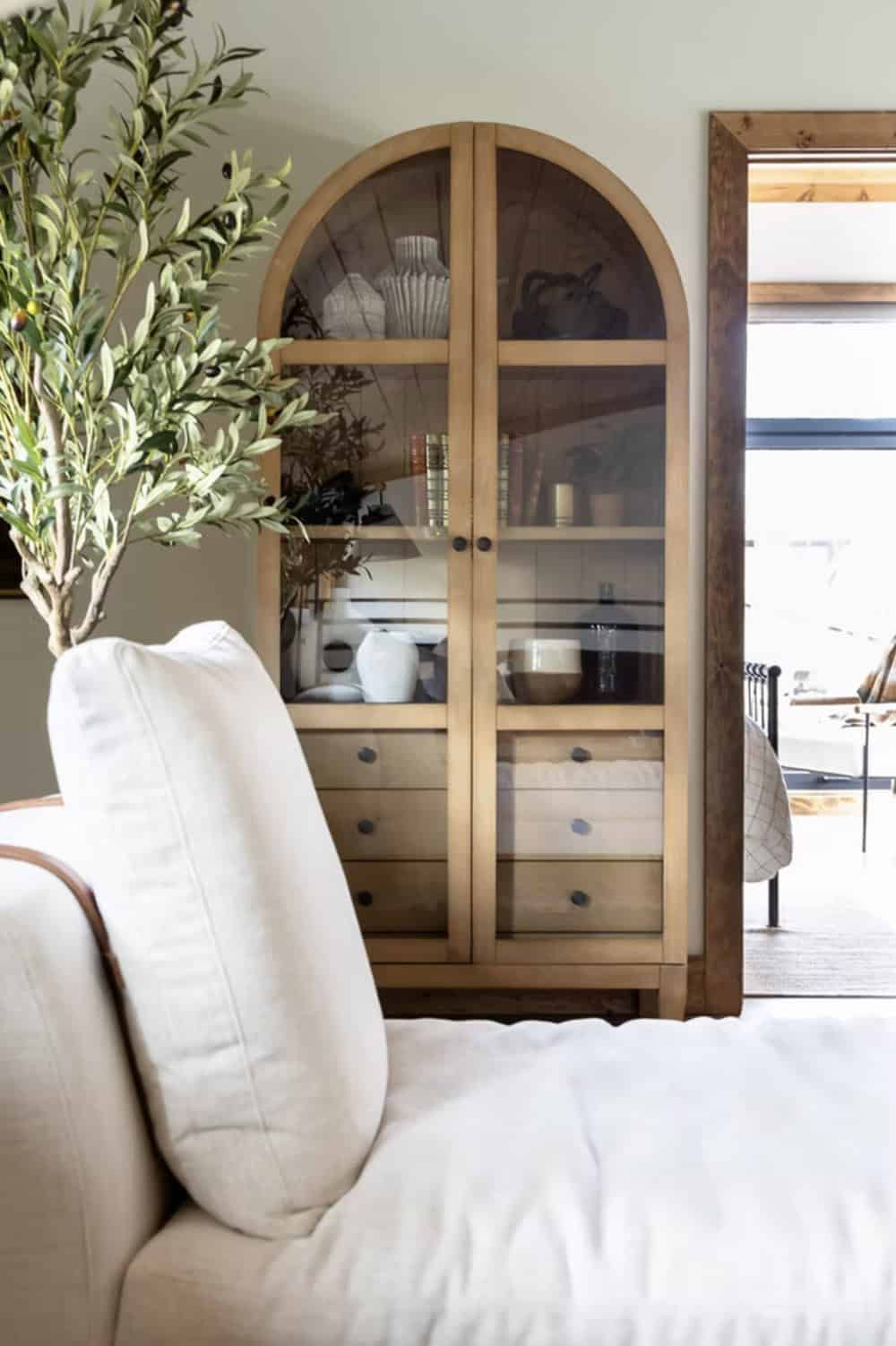 Arched wood and glass display cabinet styled with white ceramics and an olive branch