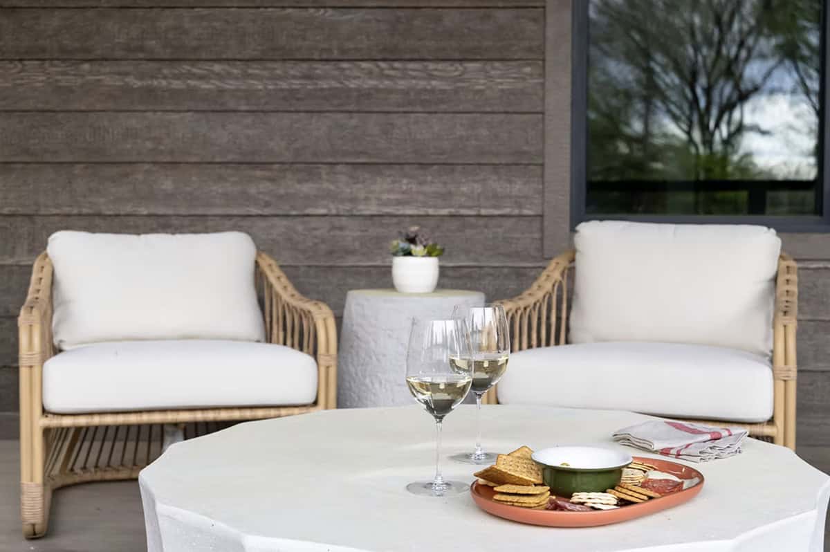 Close-up of rattan chairs with white cushions and a concrete table styled with wine and charcuterie