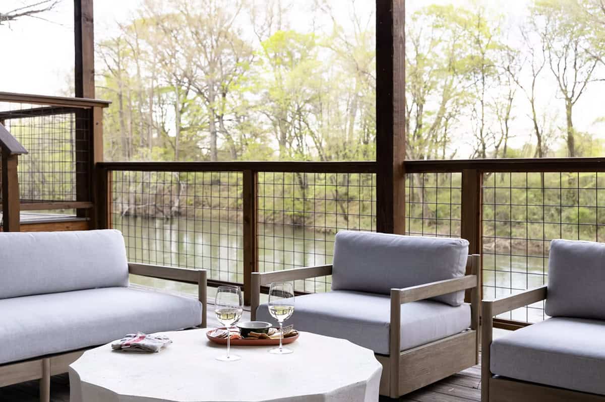 Outdoor lounge seating with gray cushions and hog-wire railing overlooking a wooded creek