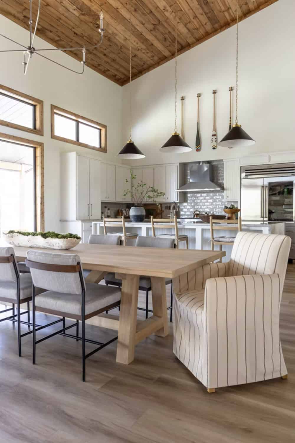 Dining area with light wood trestle table, mixed seating, black pendant lights, and white kitchen