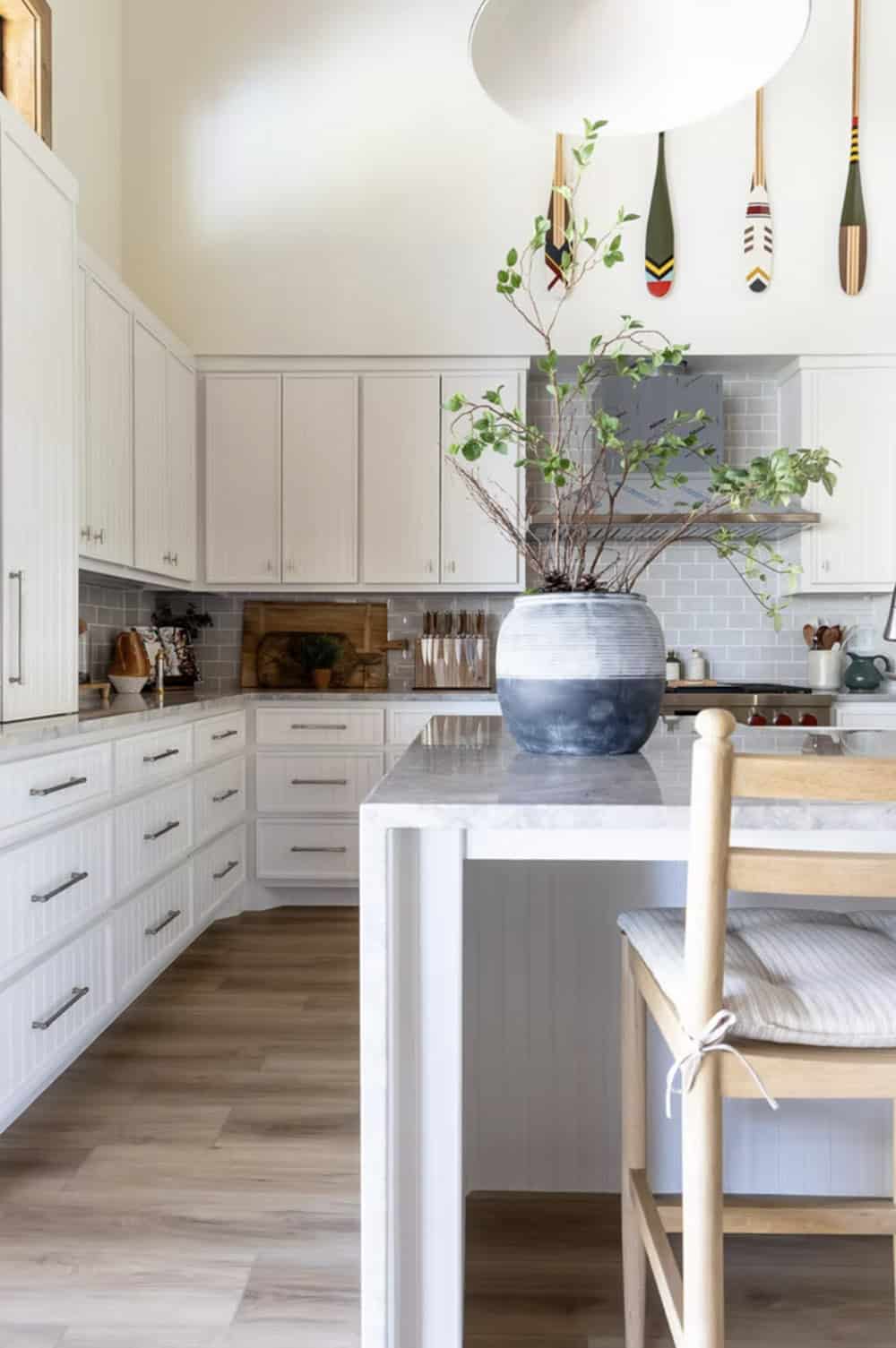 White beadboard kitchen cabinets with marble countertops, large ceramic vase, and decorative oars above