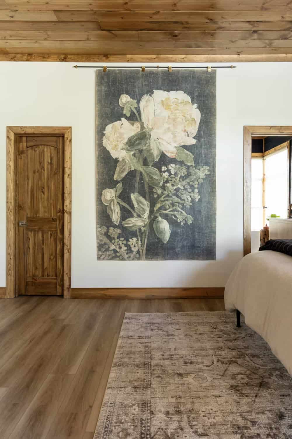 Bedroom wall with oversized botanical floral tapestry hung on brass rod, vintage rug, and wood ceiling