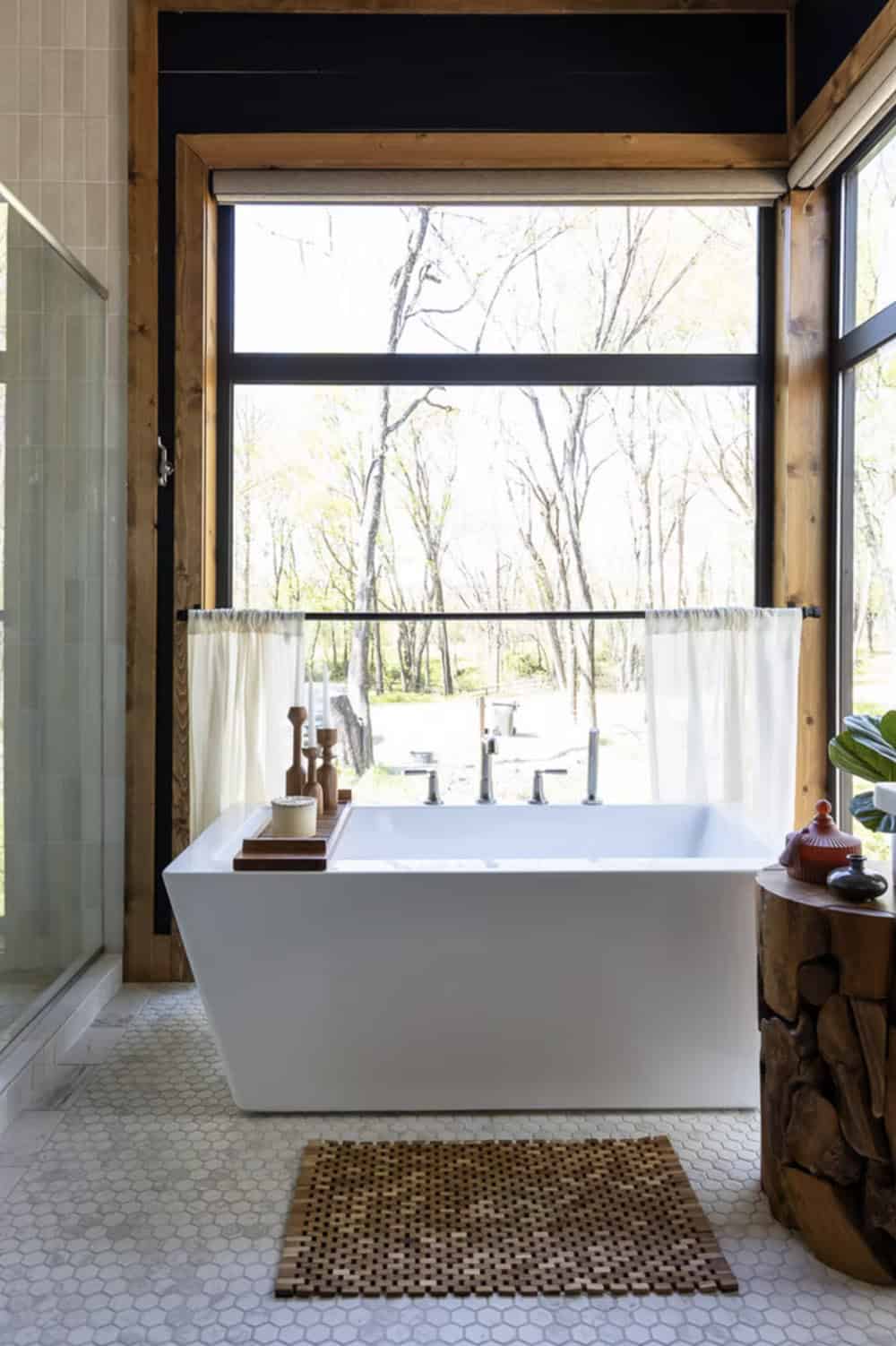 Freestanding soaking tub in front of large black-framed windows with wooded views and wood bath mat
