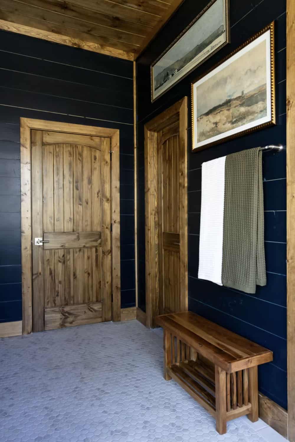 Main bathroom with dark shiplap walls, knotty wood door, teak bench, and landscape art above towel bar