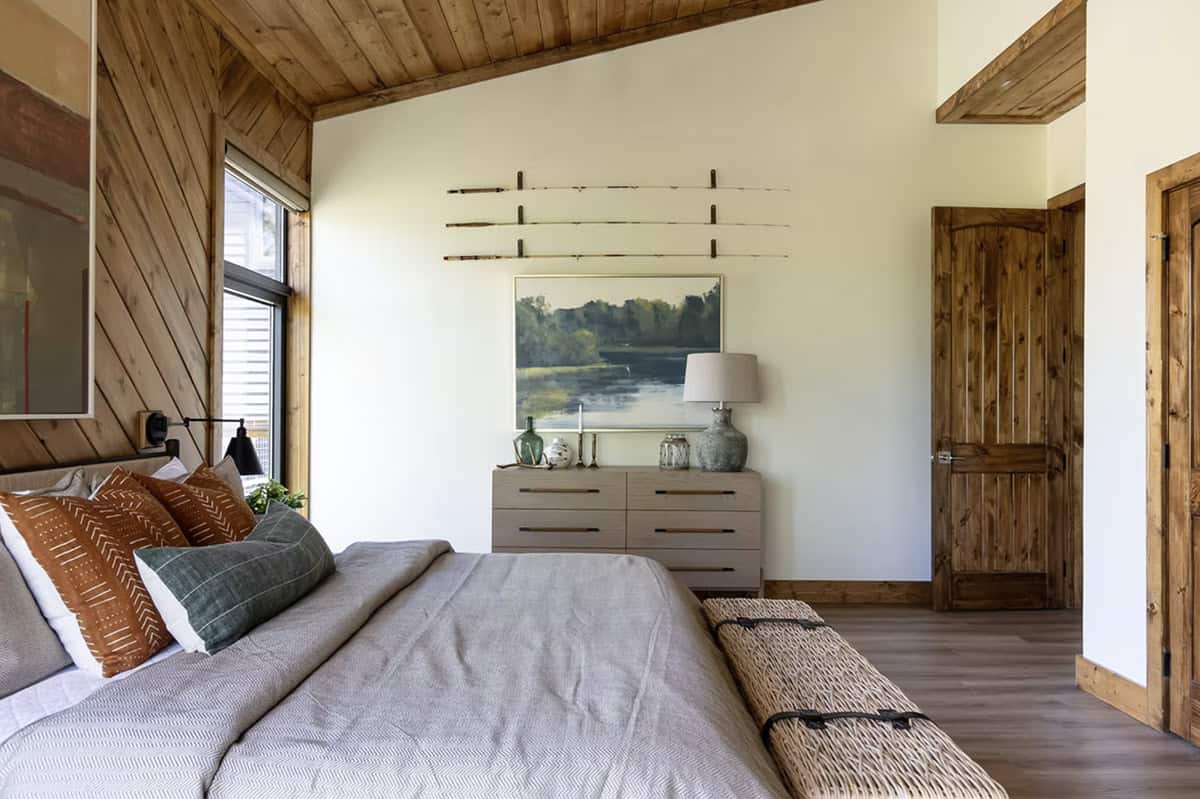 Guest bedroom with diagonal wood walls, gray linen bedding, wicker storage bench, and vintage fishing rods on wall