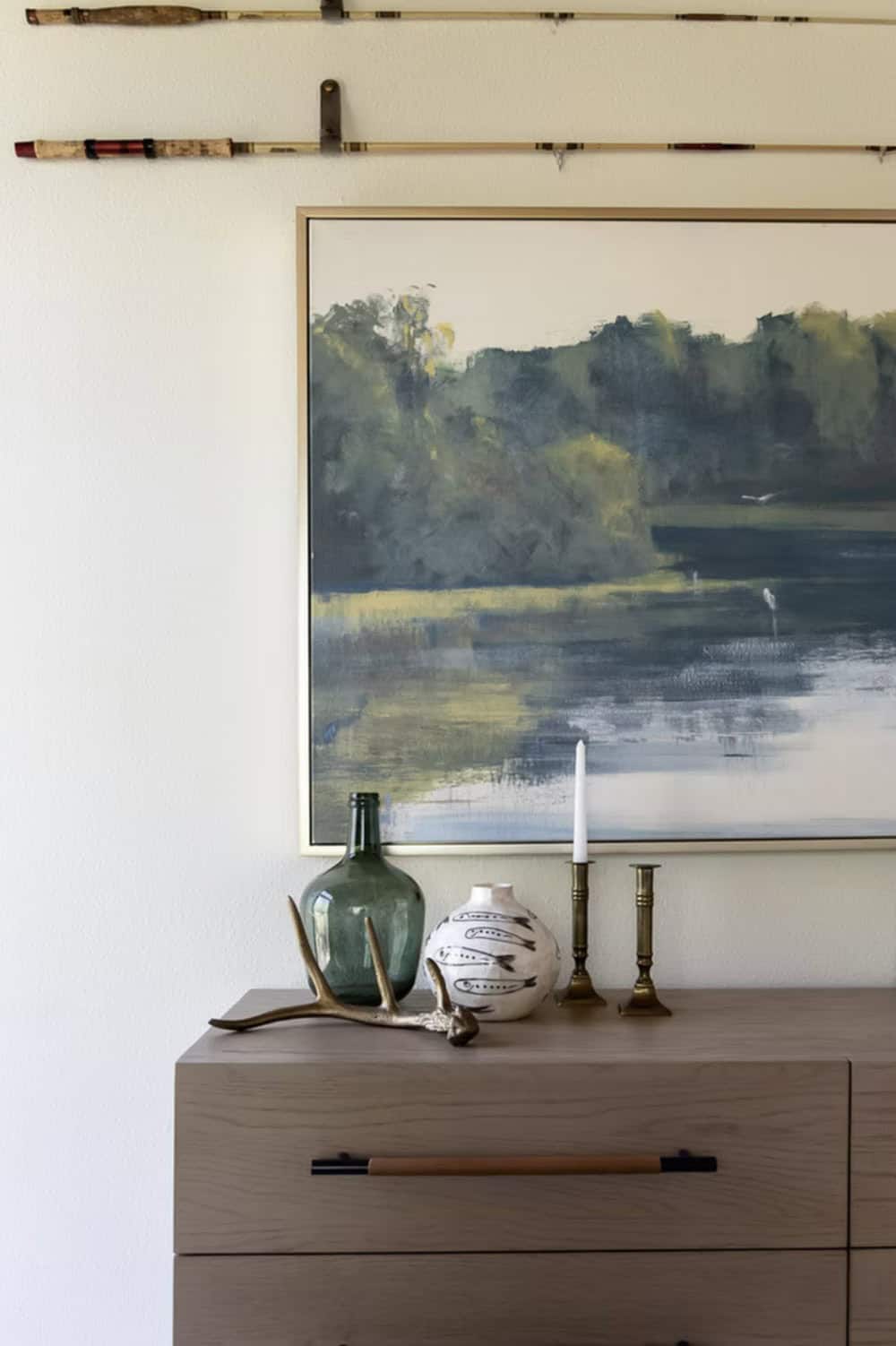 Bedroom dresser vignette with antler, green glass bottle, fish-motif vase, and brass candlestick below landscape painting