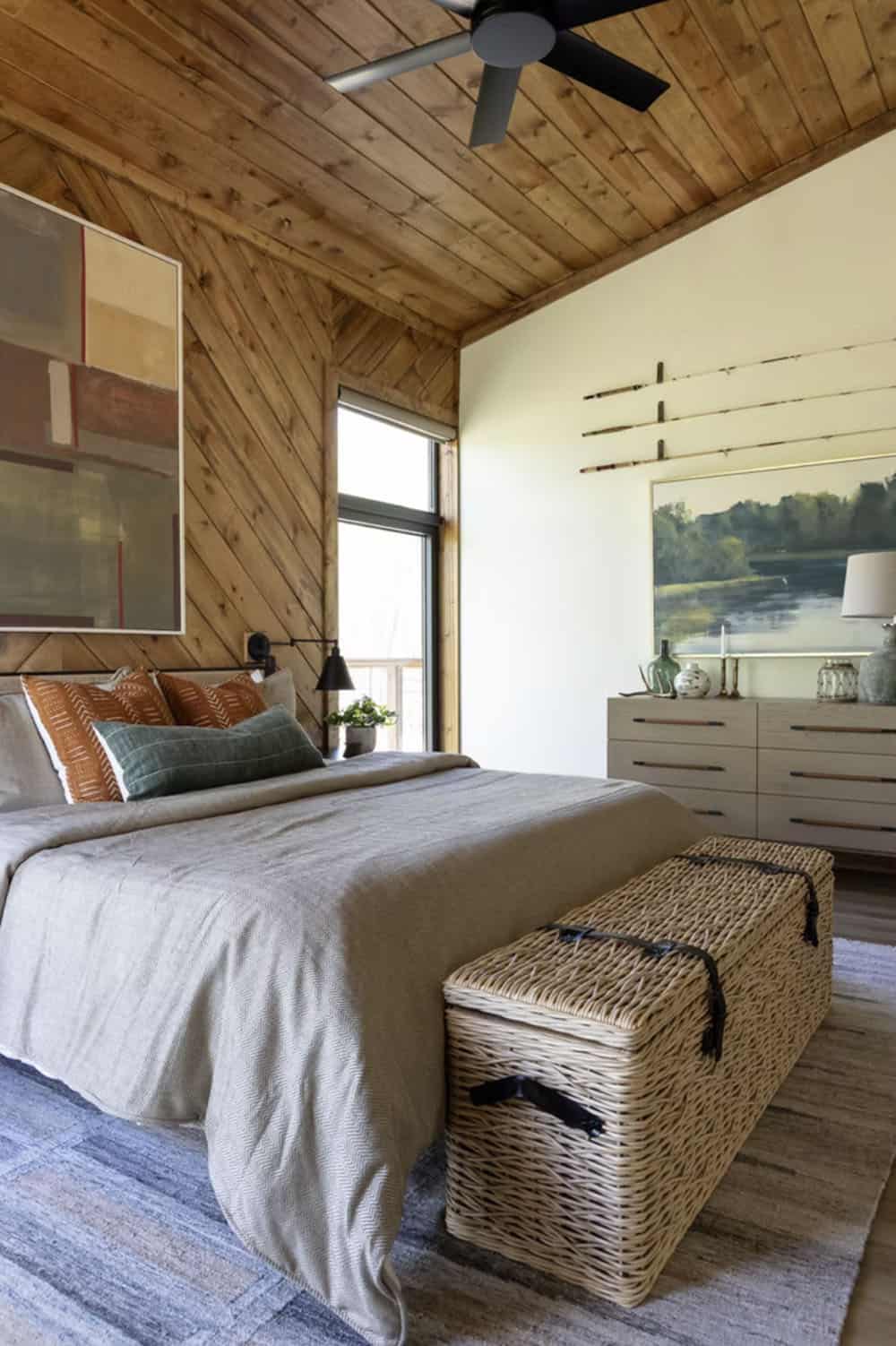 Guest bedroom with diagonal wood walls, wicker storage trunk, gray linen bedding, and lake landscape painting