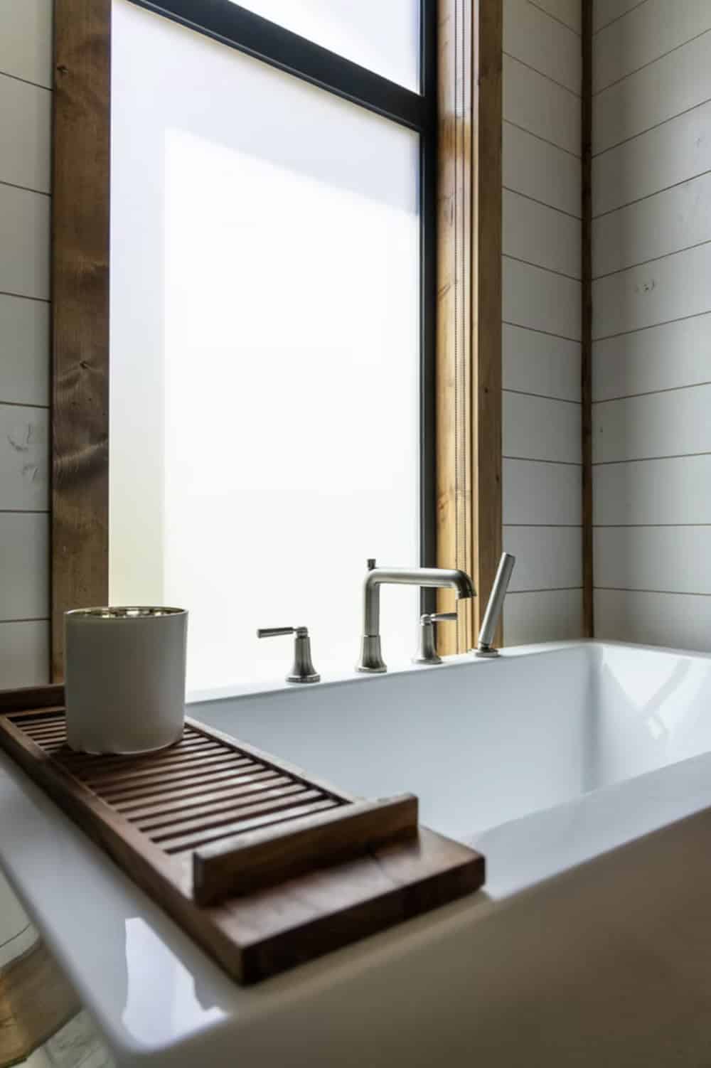Close-up of freestanding tub with wood caddy tray, ceramic cup, and brushed nickel faucet by frosted window