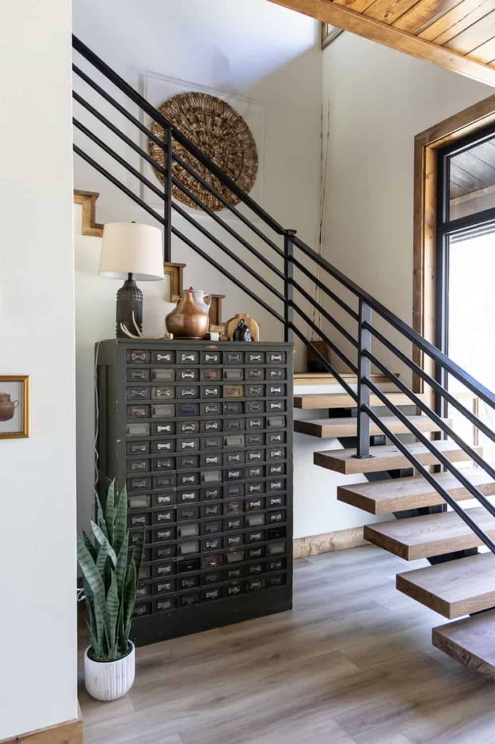 Staircase with black metal railing, floating wood treads, vintage multi-drawer cabinet, and snake plant below