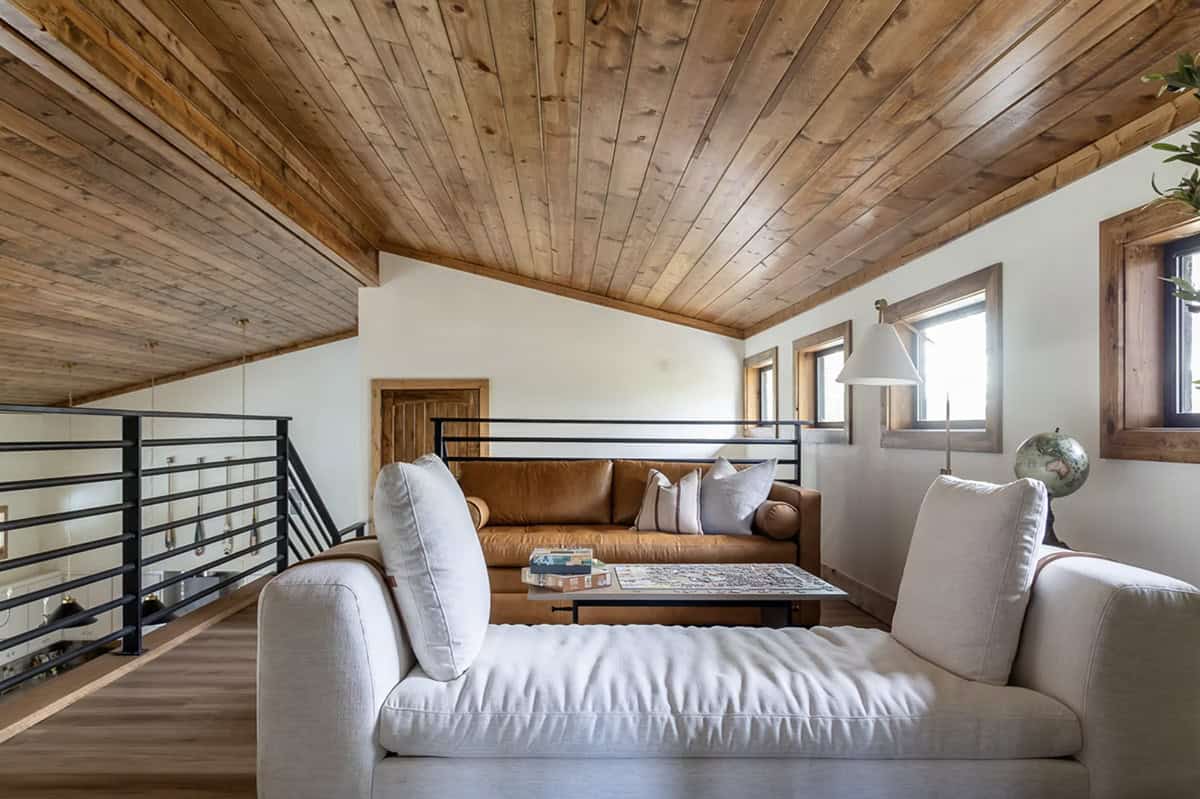 Upstairs loft lounge with tan leather sofa, white chaise, wood ceiling, and black metal railing overlooking lower level