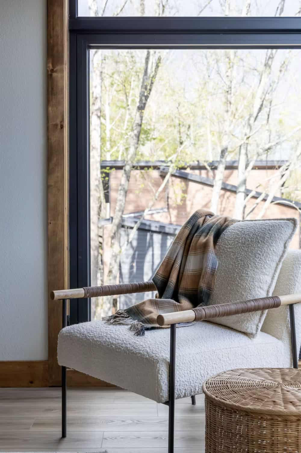 Boucle armchair with plaid throw near large black-framed window overlooking wooded cabin exterior