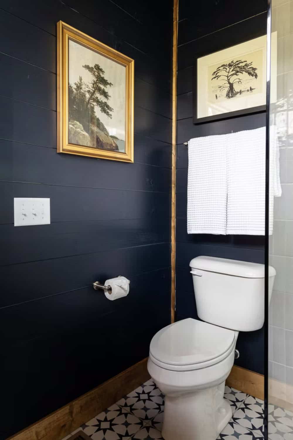 Dark navy shiplap bathroom with gold-framed landscape painting, toilet, and geometric patterned tile floor