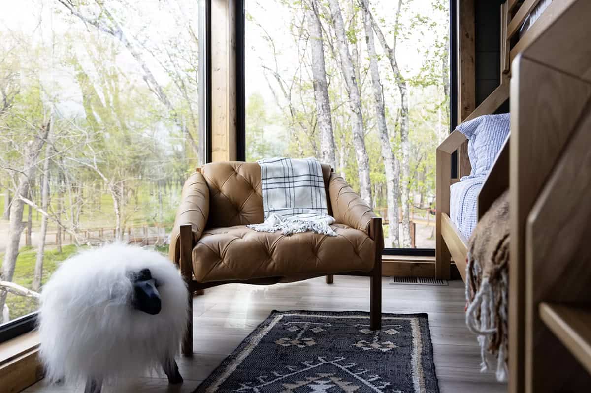 Tufted tan leather chair with plaid throw beside floor-to-ceiling windows overlooking wooded creek views