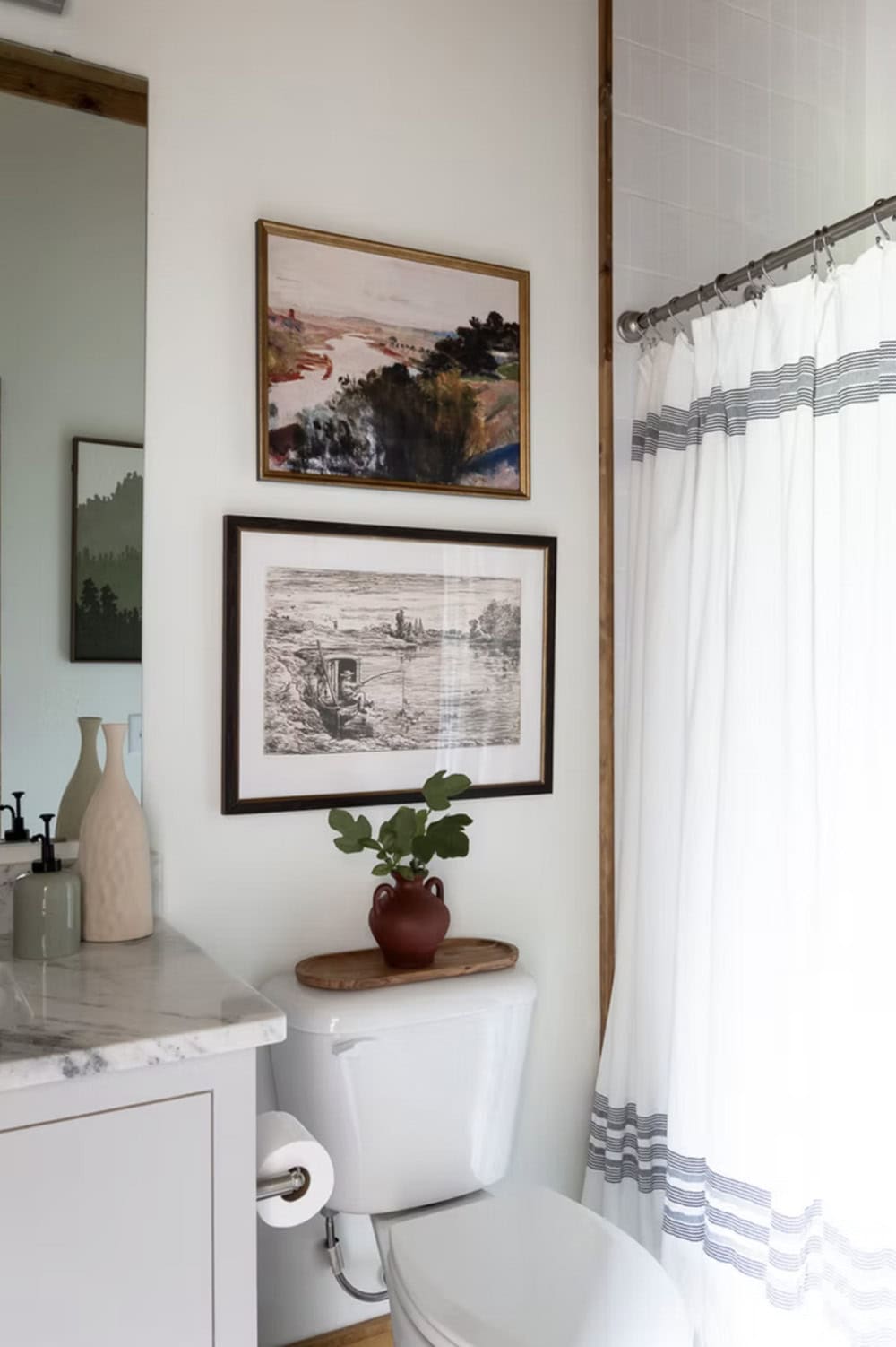 Bathroom with two landscape prints above toilet, terra cotta vase with greenery, and striped shower curtain