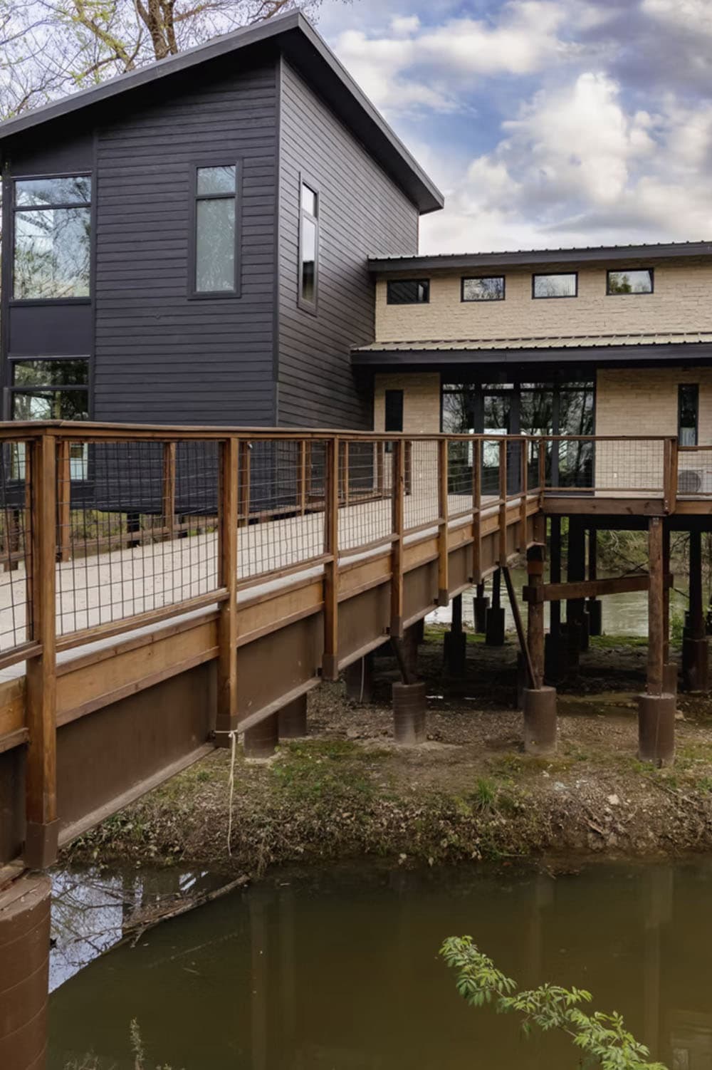 Modern rustic cabin exterior with dark siding, hog-wire railing bridge, and elevated deck over creek