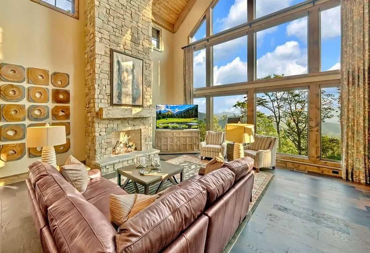 Great room with leather sectional, stone fireplace, and wall of windows overlooking mountains