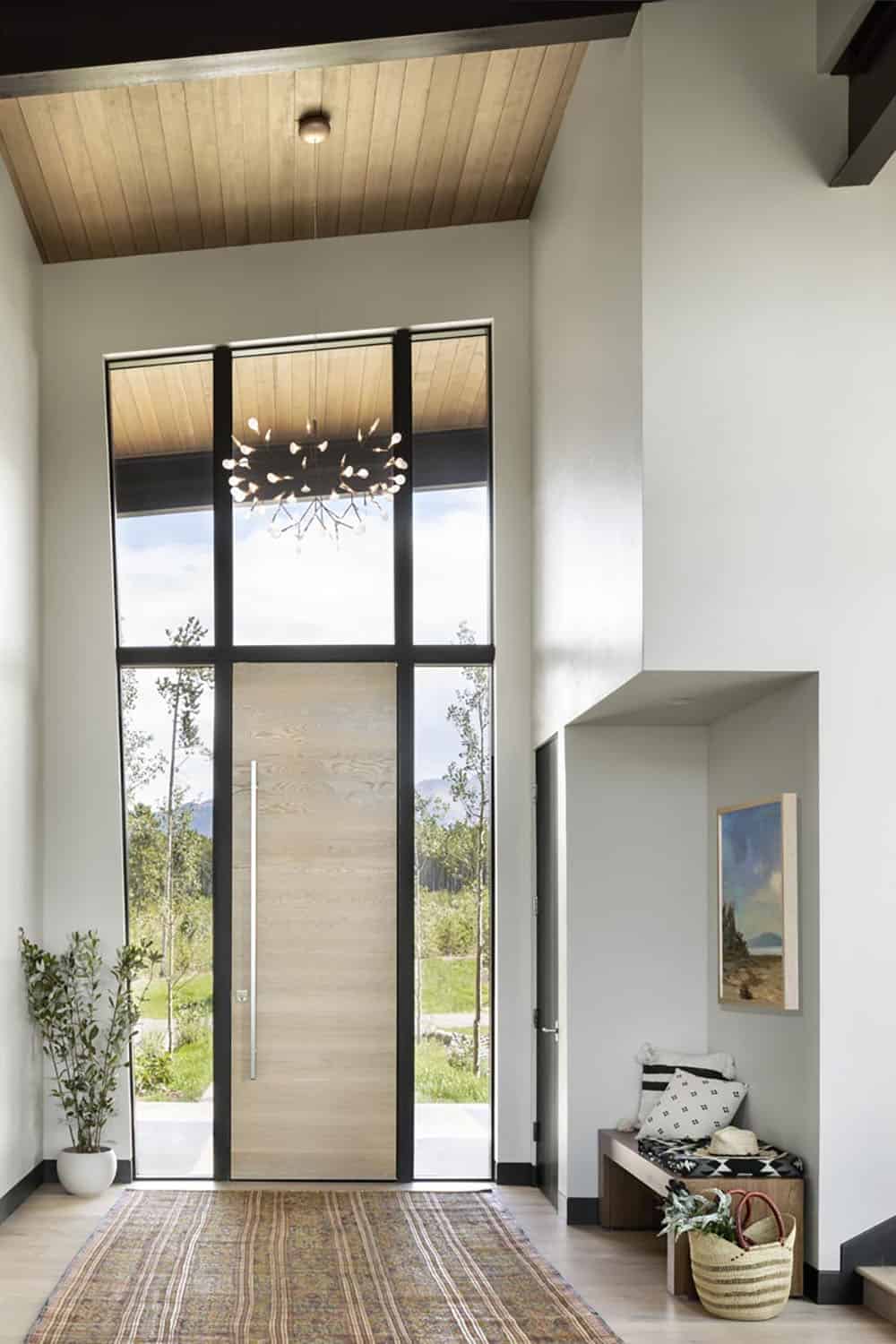 Soaring foyer with floor-to-ceiling windows, wood pivot door, and branch chandelier
