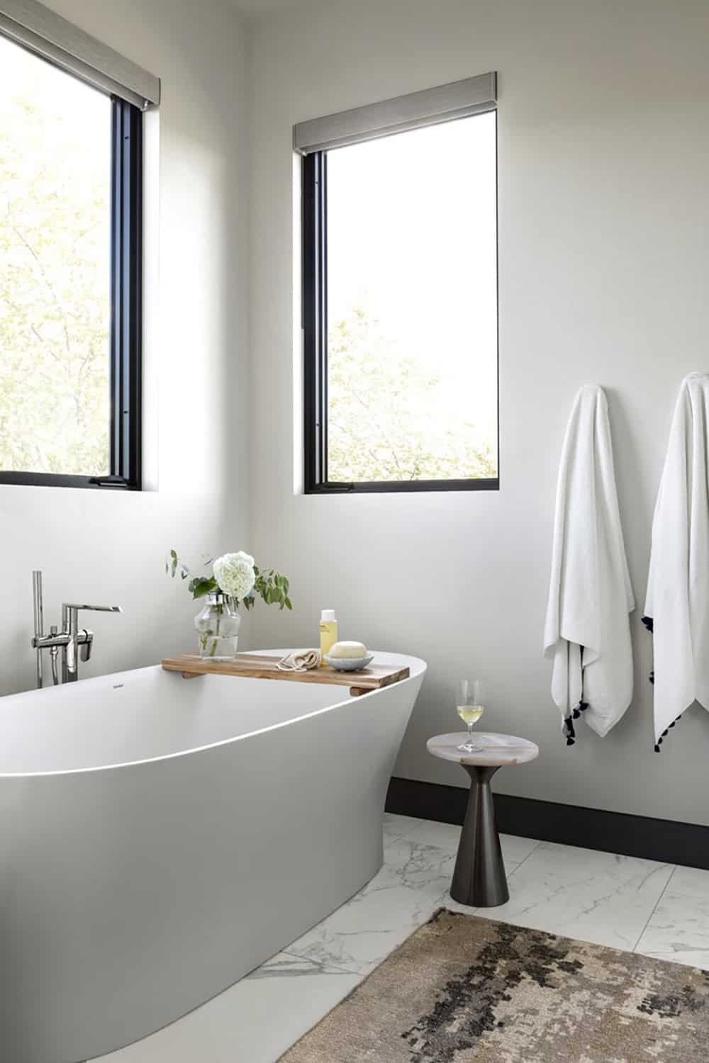 Freestanding soaking tub beneath black-framed windows with marble tile floor