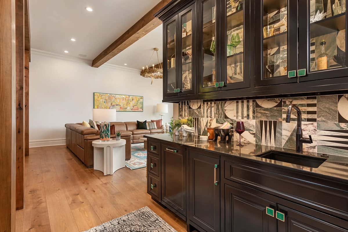 Lower level wet bar with dark cabinets, geometric stone backsplash, and open view to media room