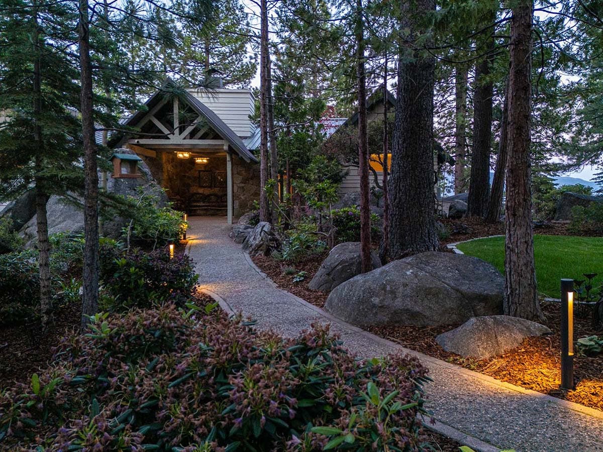 Mountain lodge exterior at dusk with illuminated pathway through pine trees and boulders